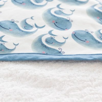 Imagen de una suave toalla de bebé doblada, con una capucha de borde azul y estampado de ballenas. Ideal para la delicada piel de tu bebé después del baño